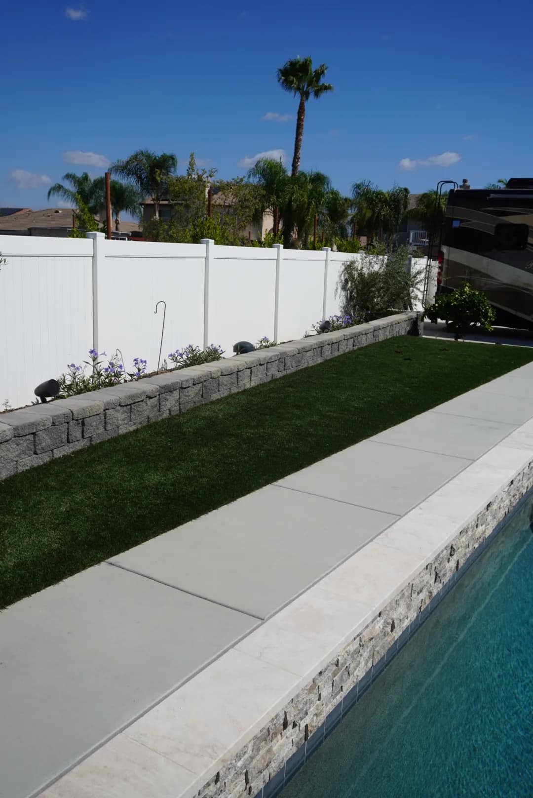 Narrow strip of fresh turf between a pool deck and a white vinyl fence
